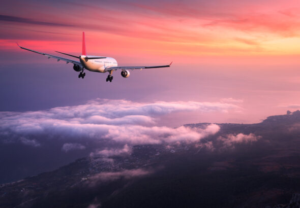 Airplane. Landscape with big white passenger airplane is flying in the red sky over the clouds at colorful sunset. Journey. Passenger airliner is landing at dusk. Business trip. Commercial plane