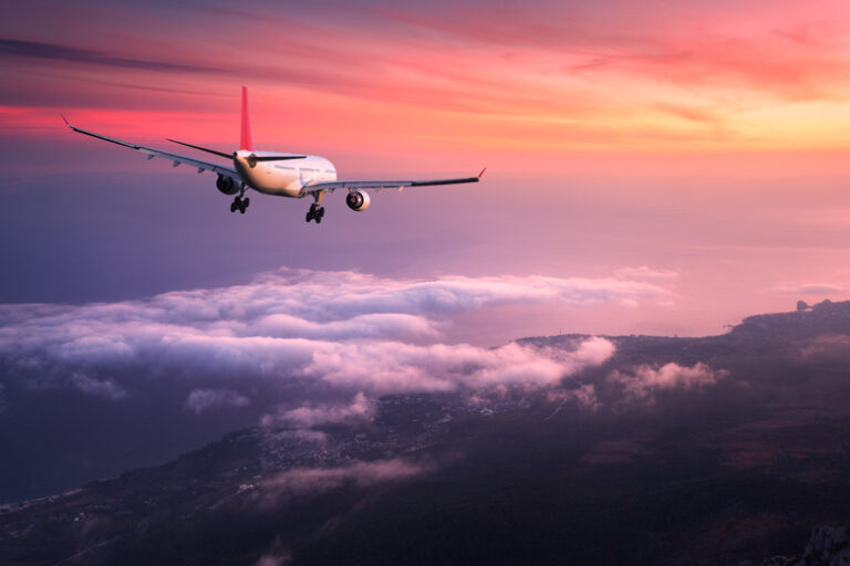 Airplane. Landscape with big white passenger airplane is flying in the red sky over the clouds at colorful sunset. Journey. Passenger airliner is landing at dusk. Business trip. Commercial plane